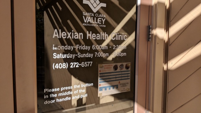 Glass entrance door displaying Alexian Health Clinic operating hours, phone contact information, and entry instructions.