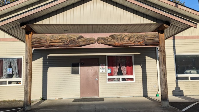 Exterior view of single-story facility building with cream siding, covered wooden entrance overhang, maroon door, and large windows.