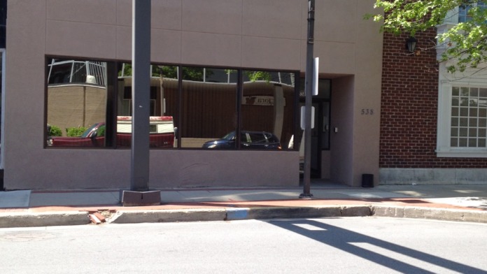 Exterior storefront with tan facade panels, dark windows, brick building, and adjacent greenery visible.