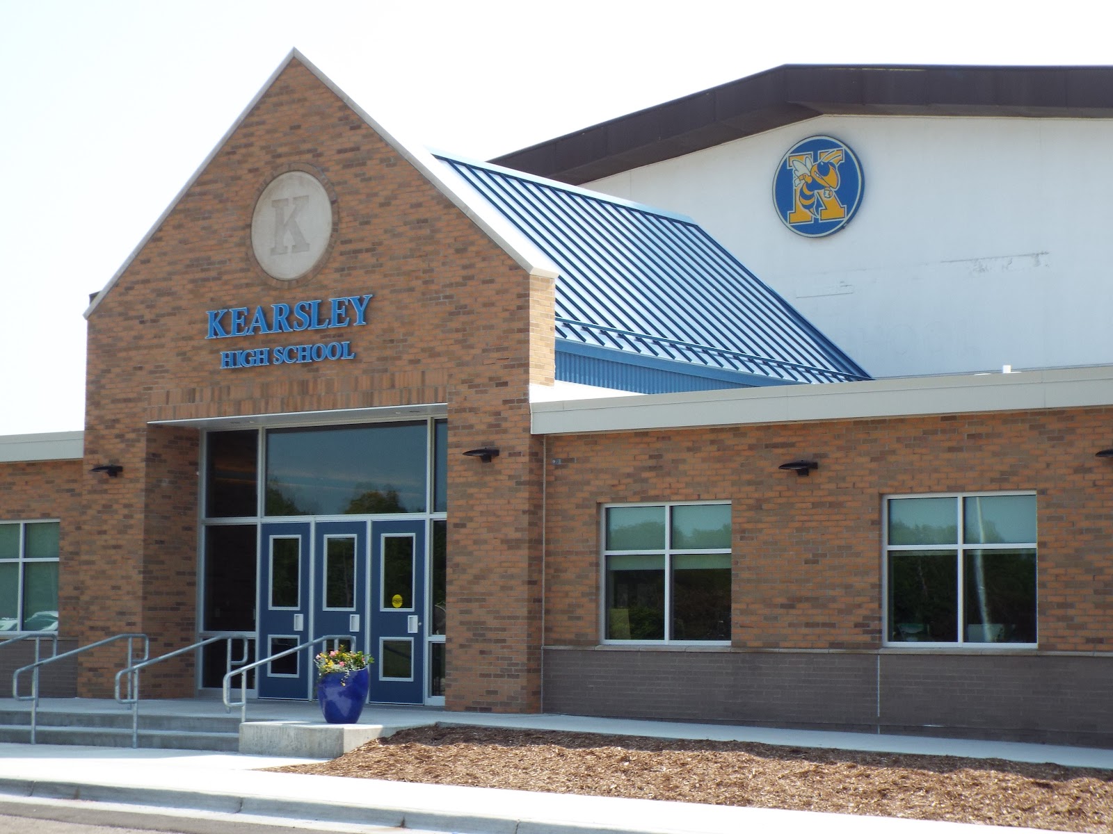 Exterior of Kearsley High School main entrance showing brick construction, blue signage, glass doors, and landscaped entry area