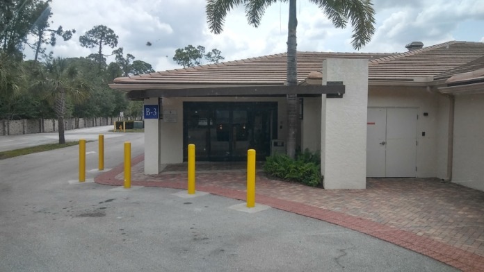 Exterior building entrance showing brick facade, protective bollards, and palm tree landscaping near main entry