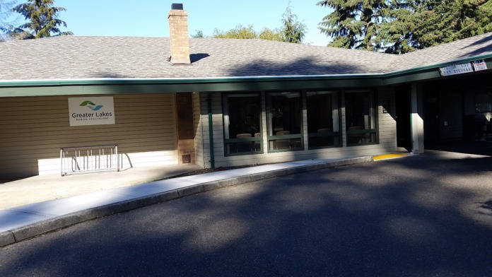 Exterior view of Greater Lakes Mental Health Spanaway clinic showing single-story building with covered entrance, signage, parking area, and surrounding evergreen trees.