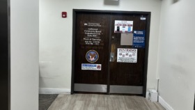 Interior hallway leading to main entrance with exit sign, white walls, wooden doors, and safety signage displayed