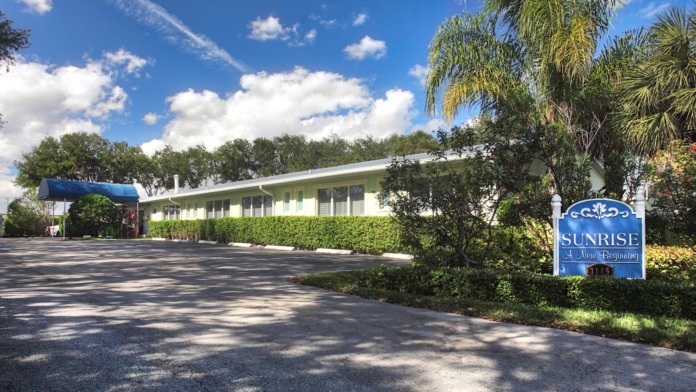 Exterior view of single-story white facility building with facility signage, tropical landscaping, and palm trees on driveway entrance approach.