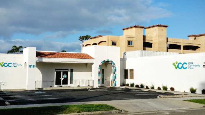 Exterior front view of Vista Community Clinic showing white building with green and blue logo, entrance with decorative tile archway, and parking area