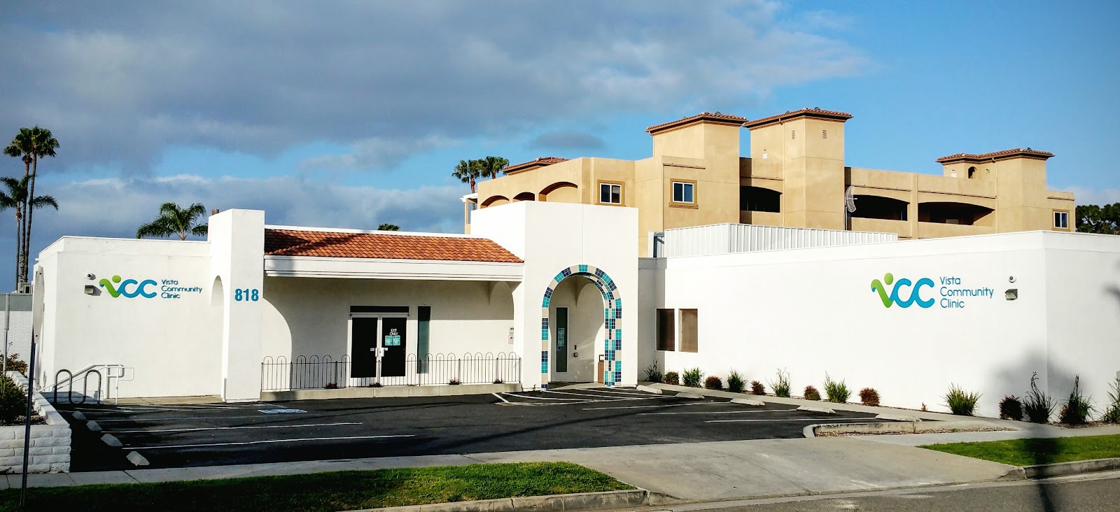 Exterior front view of Vista Community Clinic showing white building with green and blue logo, entrance with decorative tile archway, and parking area