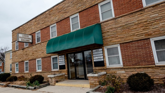Exterior front entrance of brick and wood building with green awning, multiple windows, landscaping, and entrance steps.