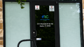 Exterior front entrance showing black framed glass doors with facility signage, address, and green ivy landscaping around stone facade
