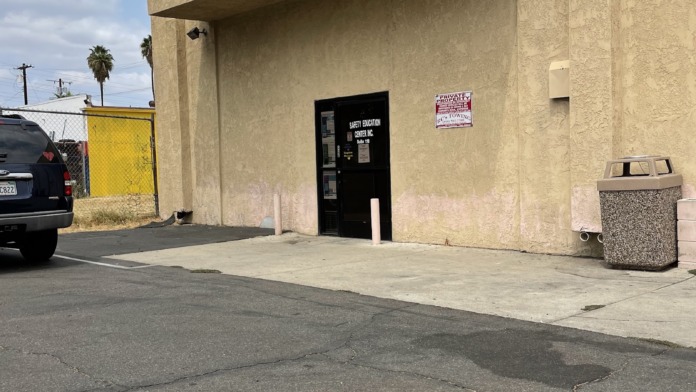 Exterior view of tan stucco building with entrance door, overhang, parking area, and utility bins.