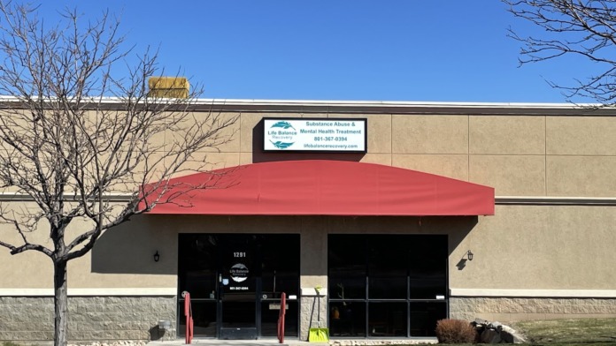 Exterior front facade with tan brick building, red awning over entrance, glass doors, and metal railings.