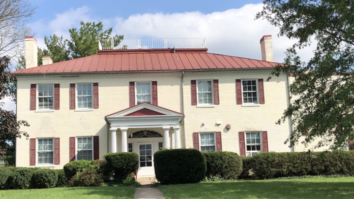 Exterior view of two-story cream-colored brick building with red metal roof, red shutters, columned portico entrance, manicured hedges, and lawn