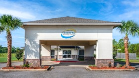 Exterior view of Oceans Behavioral Hospital main entrance with white facade, brick base, glass doors, and palm tree landscaping