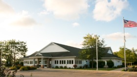 Exterior view of health center main entrance with white building, brick accents, gabled roof, landscaping, and parking area
