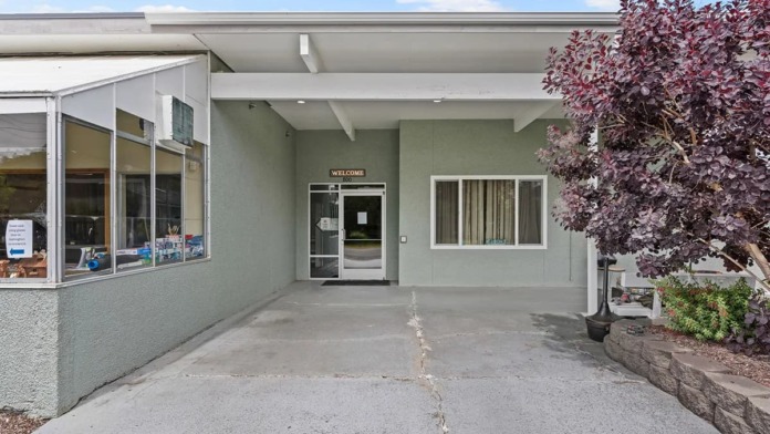 Exterior of recovery facility showing single-story building with welcoming entrance, gray facade, landscaping, and concrete walkway