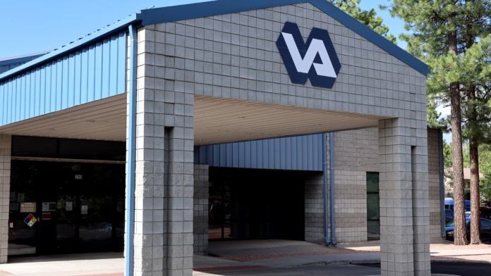 Exterior of VA health care facility showing main entrance with VA logo, stone facade, metal roofing, and parking area