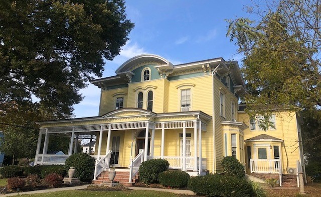 Exterior of yellow Victorian-style building with white porch columns, arched windows, and manicured landscaping