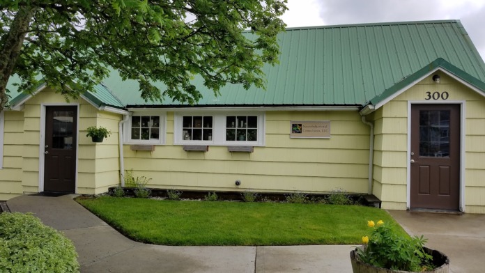 Exterior view of single-story yellow treatment facility with green metal roof, two brown doors, multiple windows, landscaped grounds, and address number 300
