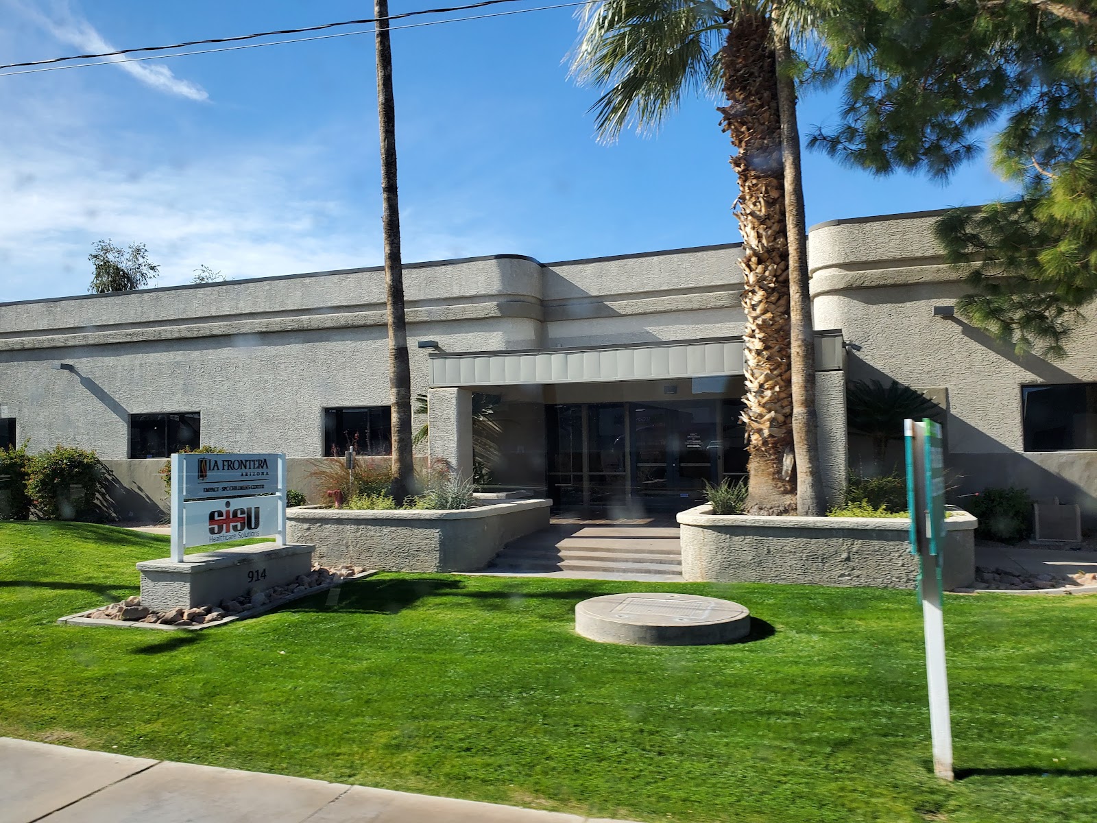 Exterior front entrance with manicured lawn, palm trees, stone signage, and facility entrance doors