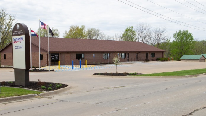 Exterior view of single-story brick medical clinic with flagpoles, accessible parking spaces, and landscaped grounds from street level.