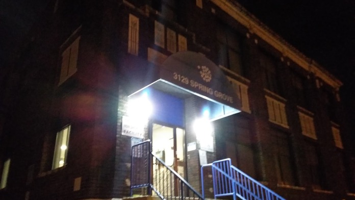 Exterior view of brick building entrance with illuminated entryway, metal railings, and stairs leading to main door at night