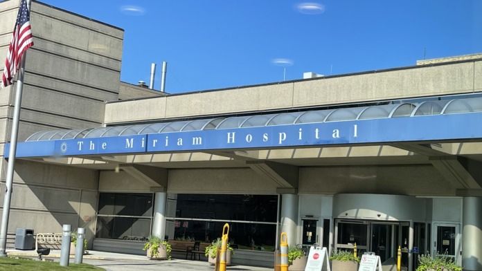 Exterior front entrance of The Miriam Hospital showing blue signage canopy, glass doors, landscaping, and American flag