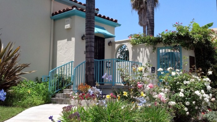 Exterior view of treatment center entrance featuring turquoise accents, flowering gardens, palm trees, and welcoming courtyard with accessible ramp