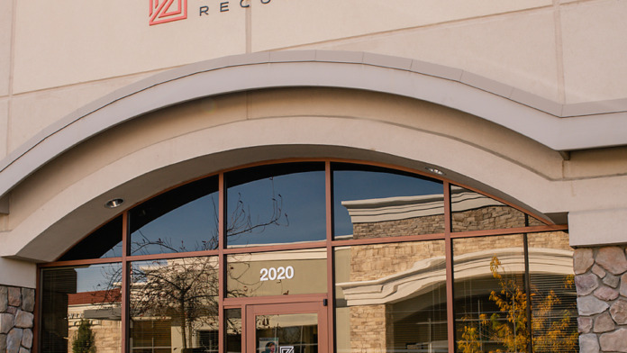 Exterior view of treatment facility main entrance showing stone cladding, Zelus Recovery signage, glass doors with brown framing, and landscaped grounds with ornamental grasses