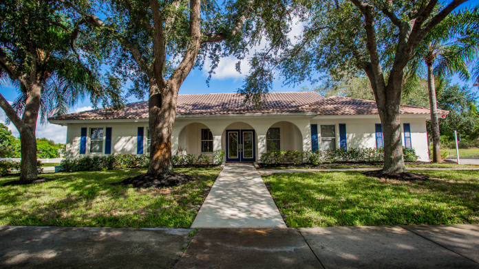 Exterior front view of single-story white treatment facility with tile roof, blue-trimmed windows, mature trees, and manicured grounds.