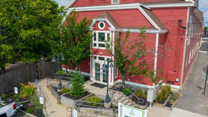 Exterior view of red historic building with white trim, entrance with railings, landscaping, and Pine Tree signage