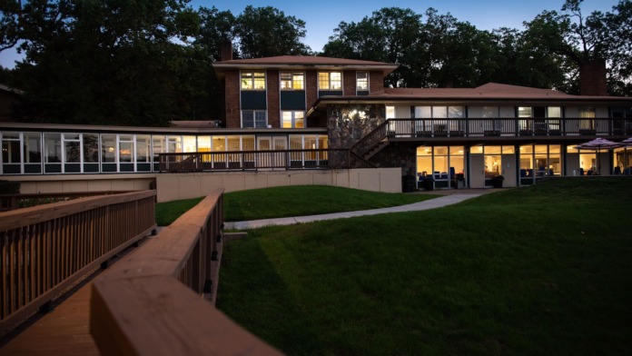 Exterior view of modern treatment facility building with lit windows, wooden railings, and lawn at evening