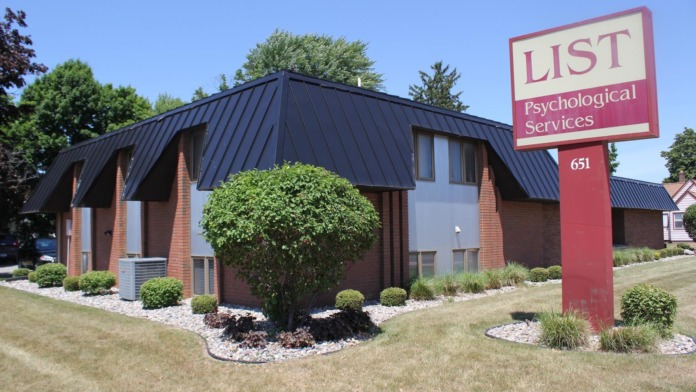 Exterior of brick psychological services building with black metal roof, landscaping, and facility sign displaying address 651