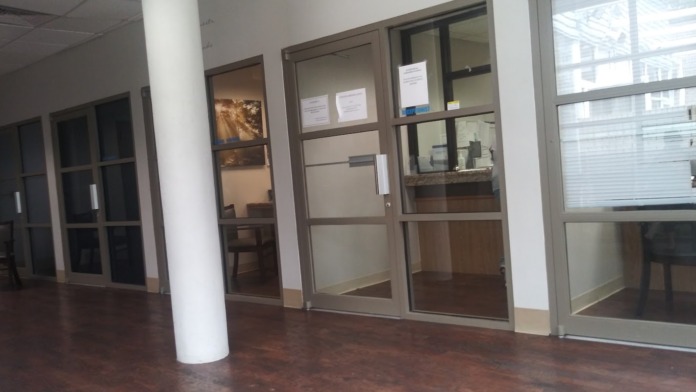 Interior hallway with glass-fronted office doors, white support columns, brown flooring, and framed artwork on walls