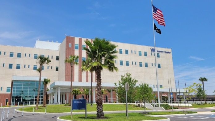Exterior front of hospital with beige and brick facade, glass entrance, windows, flags, and palm landscaping