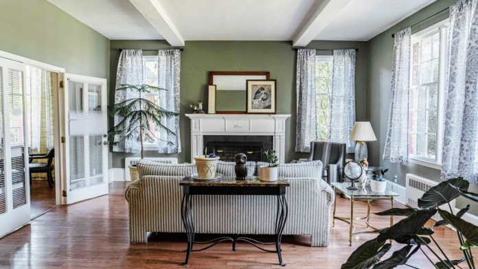 Interior living room with green walls, fireplace, striped seating, multiple windows with curtains, and decorative plants