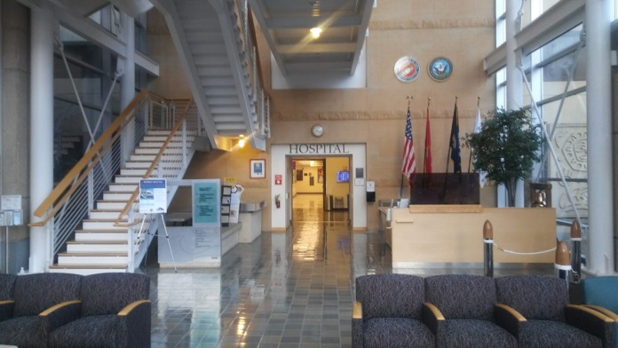 Interior hospital lobby featuring seating areas, staircase, military flags, reception desk, and signage