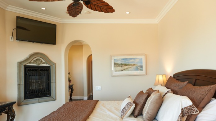 Interior spacious bedroom with king bed, decorative fireplace, flat-screen TV, tropical ceiling fan, and neutral color palette