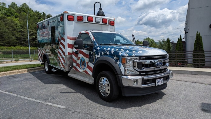 Exterior ambulance vehicle with red cross emblem and patriotic American flag livery parked on facility grounds near building entrance