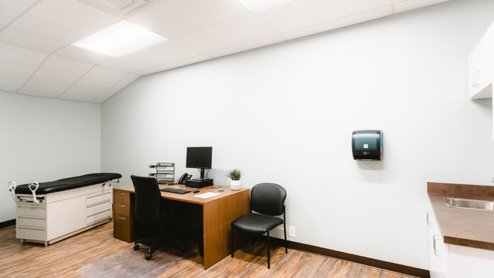 Medical office with wooden desk, computer setup, examination table, and professional office furniture and cabinetry.
