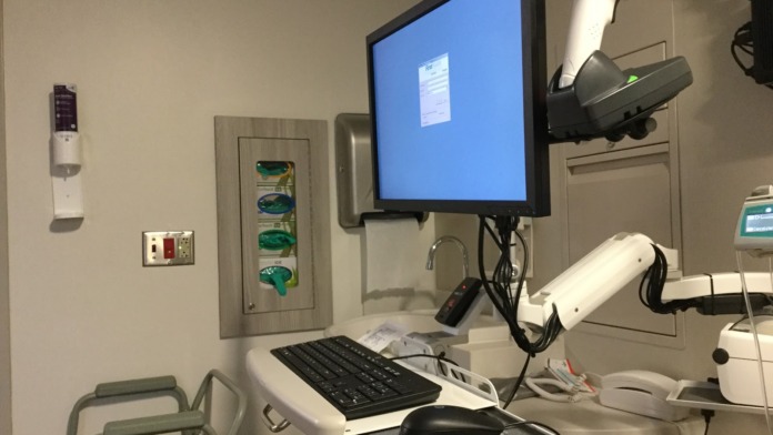 Interior clinical room with desktop computer monitor, keyboard, mouse pad, wall-mounted medical instrument organizer, and diagnostic equipment fixtures