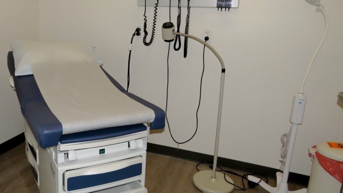 Interior clinical exam room with examination chair, wall-mounted vital signs monitor, blood pressure cuff, and medical equipment