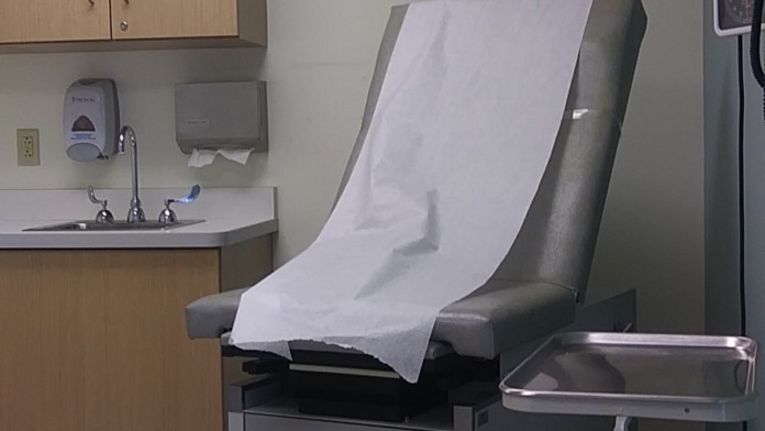 Interior patient exam room with examination table, sink, wood cabinetry, wall-mounted TV, and medical supplies storage
