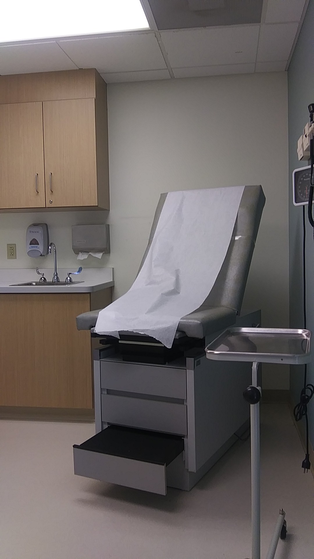 Interior patient exam room with examination table, sink, wood cabinetry, wall-mounted TV, and medical supplies storage