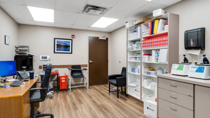 Interior clinical office with desk, computer monitor, medical equipment storage, and supply shelving