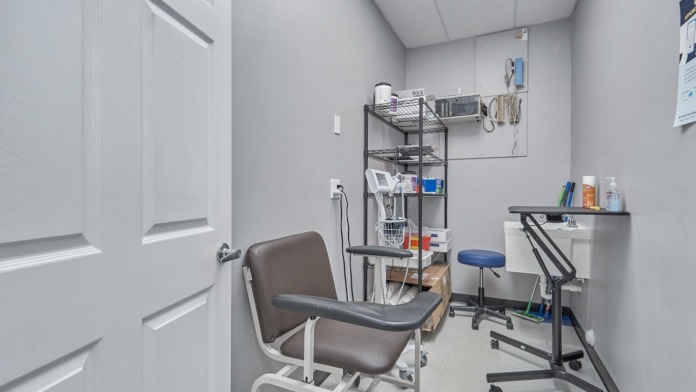 Interior clinical treatment room with examination chair, work surface, shelving with supplies, and medical equipment