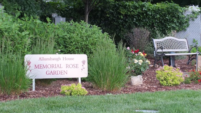 Outdoor memorial rose garden with seating benches, flowering plants, ornamental grasses, trees, and designated sign area
