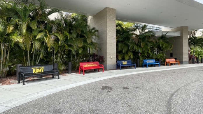 Exterior covered walkway with benches honoring military service branches, palm trees, and concrete pathway