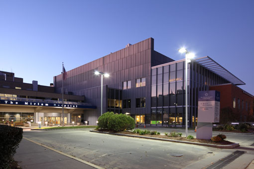 Exterior view of modern facility building with glass and dark metal facade, illuminated at dusk with landscaping and parking area
