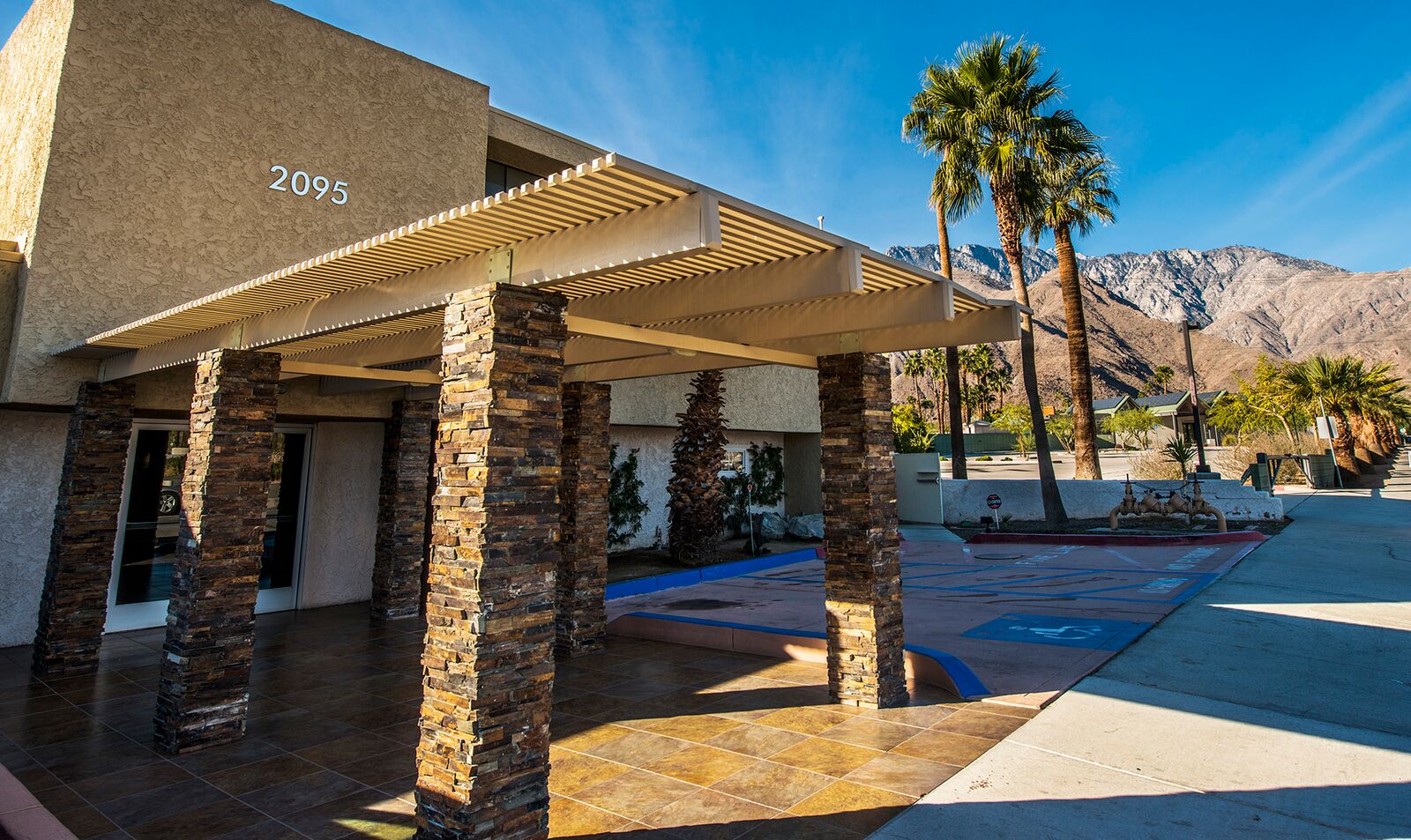 Exterior patio area with modern pergola design, stone support columns, address number 2095, metal railing, and palm trees visible
