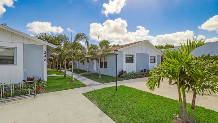 Exterior view of multiple white residential-style buildings connected by covered walkways, landscaped grounds with palms and flowering plants.