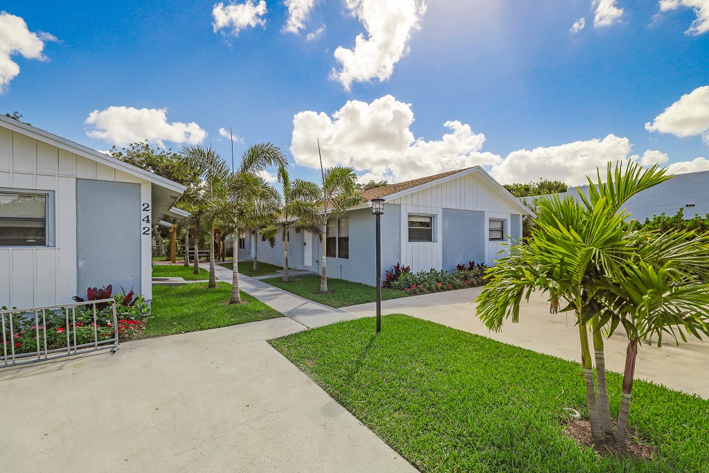 Exterior view of multiple white residential-style buildings connected by covered walkways, landscaped grounds with palms and flowering plants.
